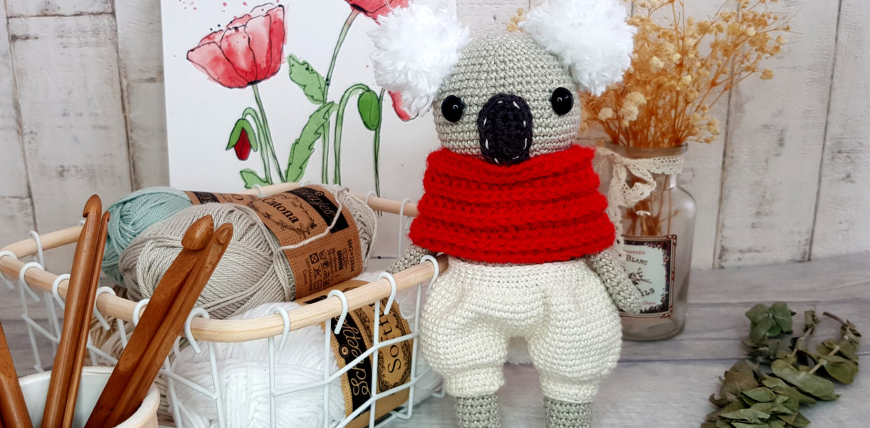 crochet koala with red scarf and white shorts standing next to a basket full of yarns