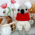 crochet koala with red scarf and white shorts standing next to a basket full of yarns