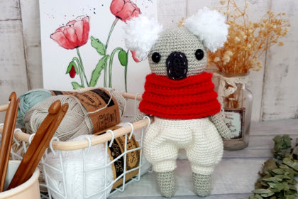 crochet koala with red scarf and white shorts standing next to a basket full of yarns