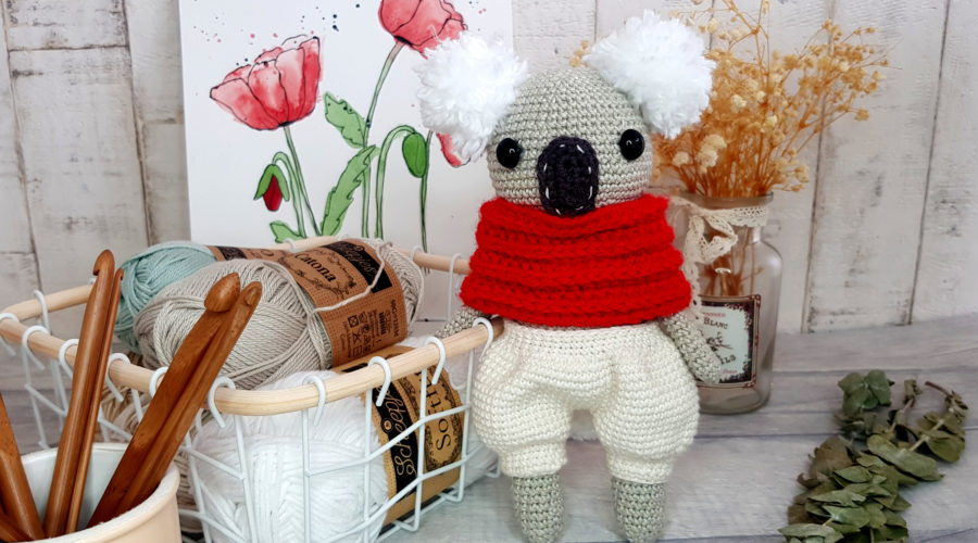 crochet koala with red scarf and white shorts standing next to a basket full of yarns