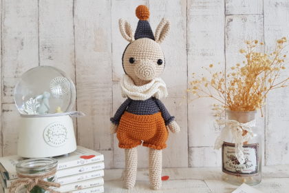 crochet llama standing next to a music box. the llama is wearing a hat, a pair of trousers and three scarves