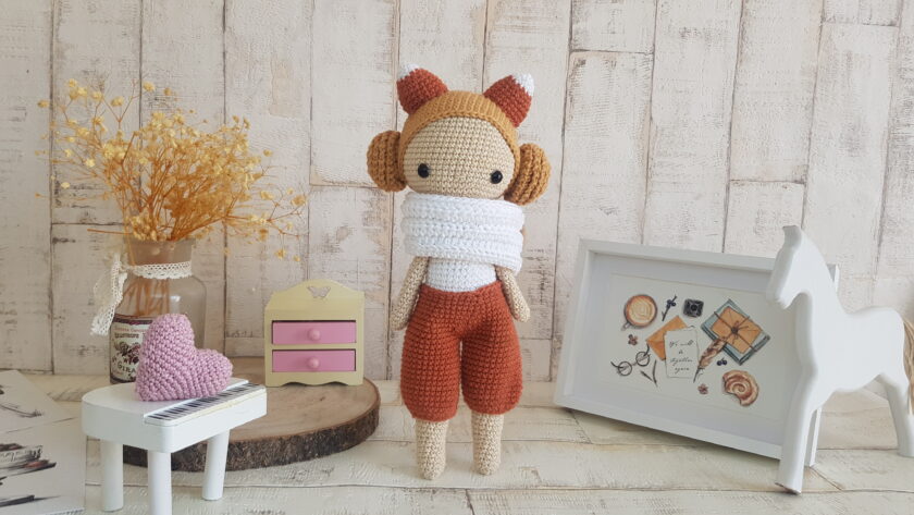 crochet doll with fox ears standing next toa piano, a rocking horse and a nice painting